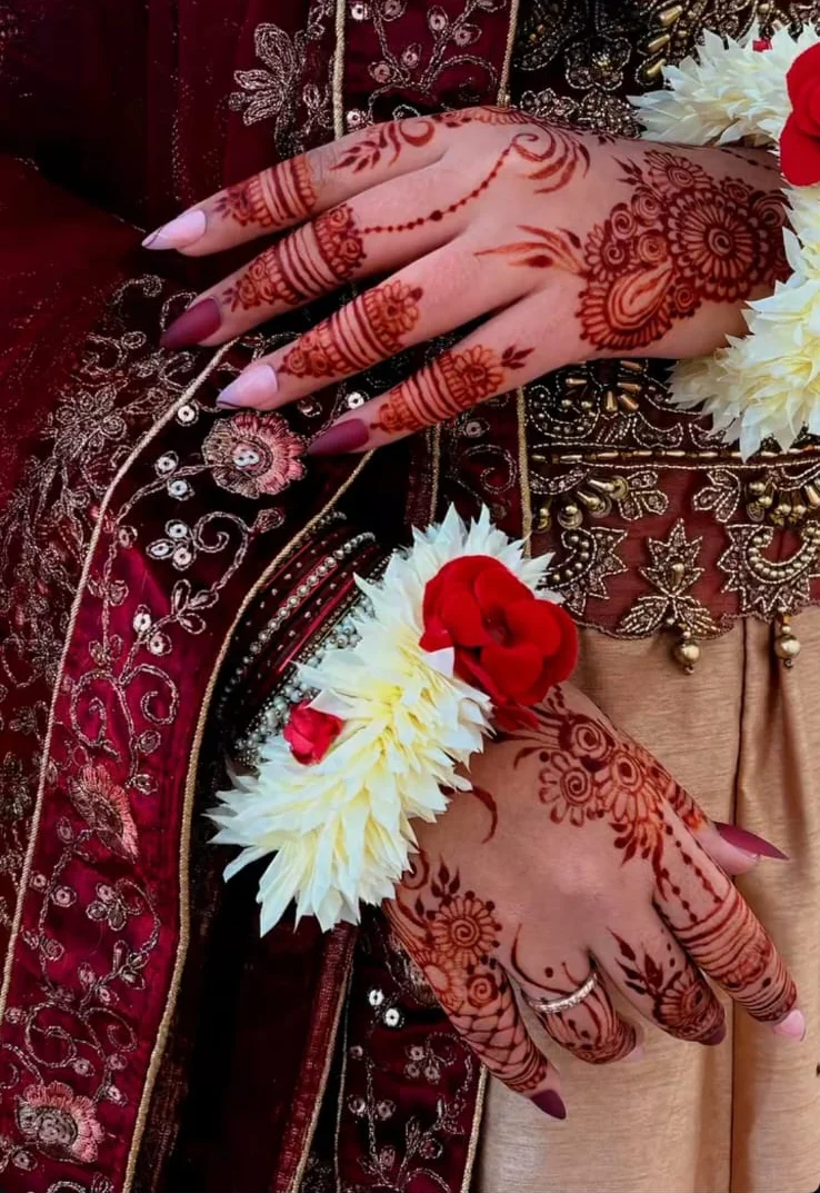 Floral and paisley mehndi on two hands against burgundy embroidery, with rose and jasmine wrist flowers